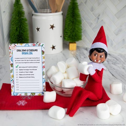 an elf with marshmallows in front of a recipe board on a kitchen counter