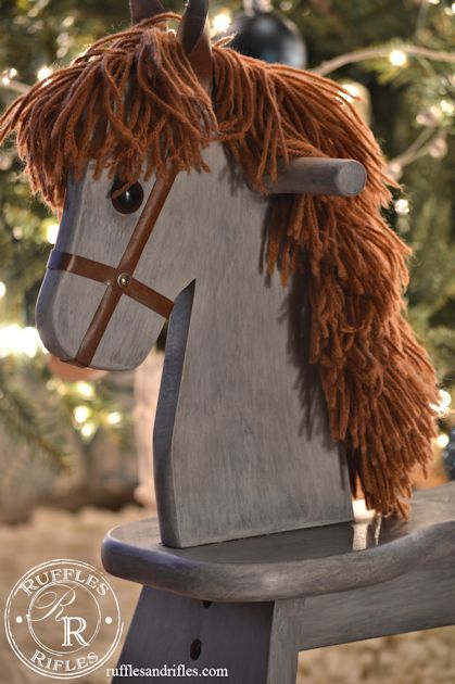 a wooden rocking horse with long hair on it's head and nose, sitting in front of a christmas tree