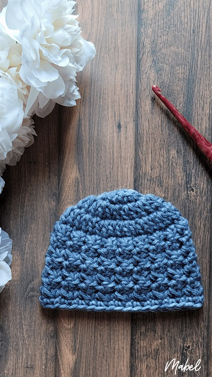 a crocheted hat sits next to some flowers on a wooden table with a pair of scissors