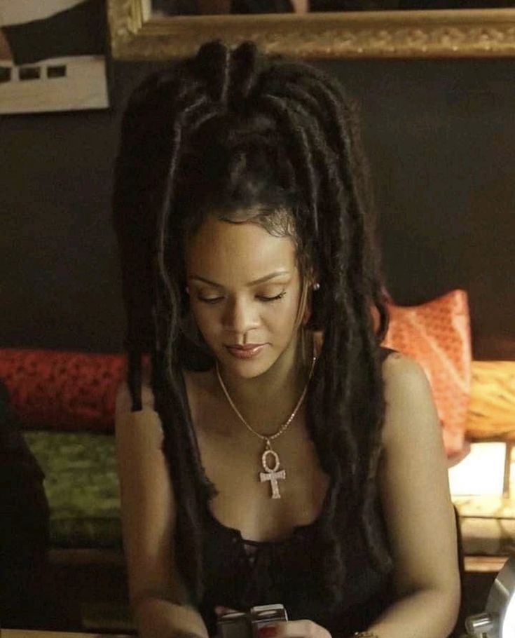 a woman with dreadlocks sitting at a table looking at her cell phone and holding a cup