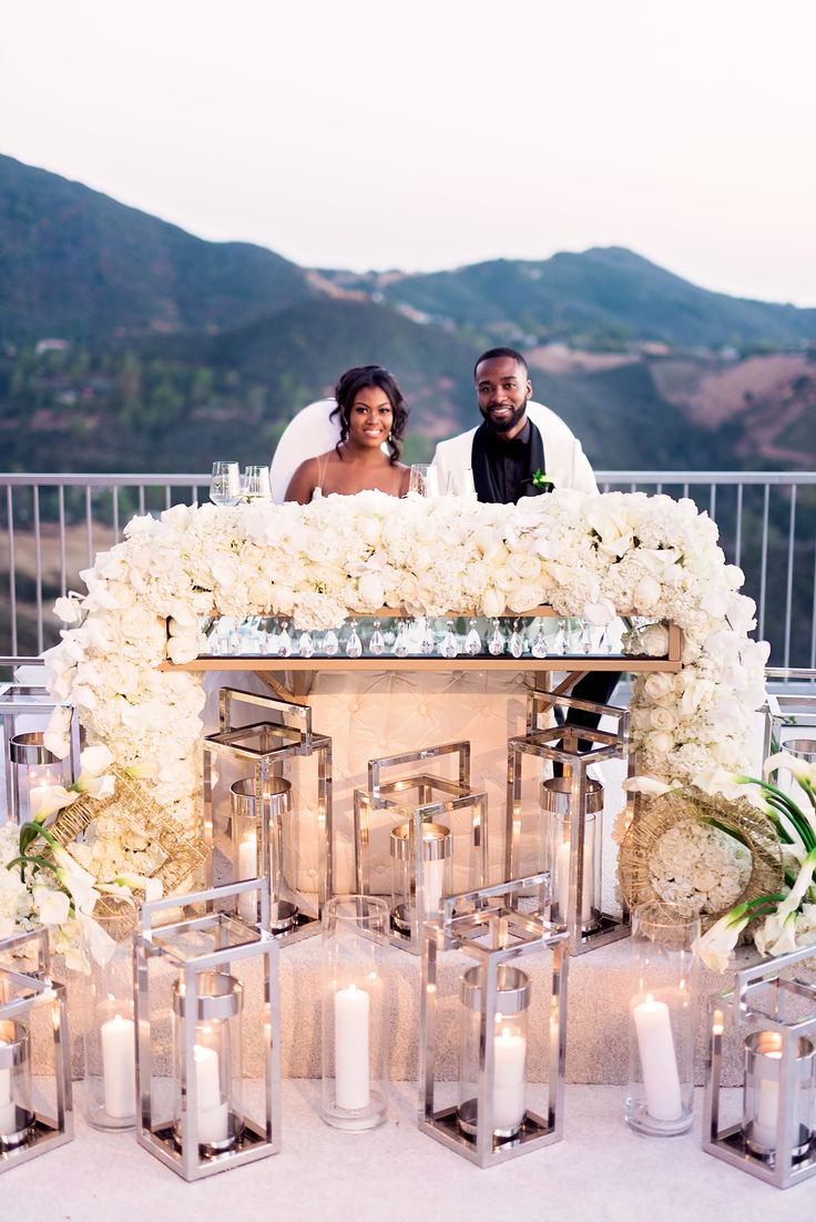 a bride and groom are sitting on a table with candles in front of them, surrounded by white flowers