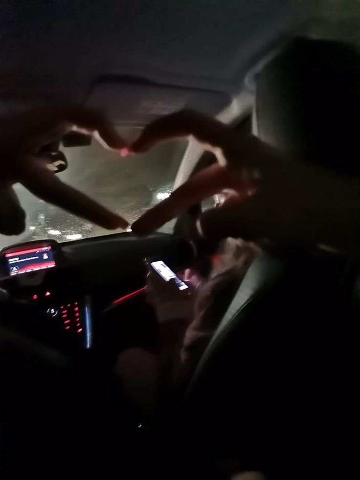 two people sitting in a car with their hands on the steering wheel, and one person using a cell phone