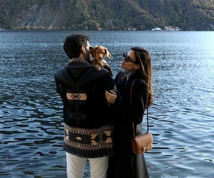 a man and woman standing next to each other with a dog on their lap looking at the water