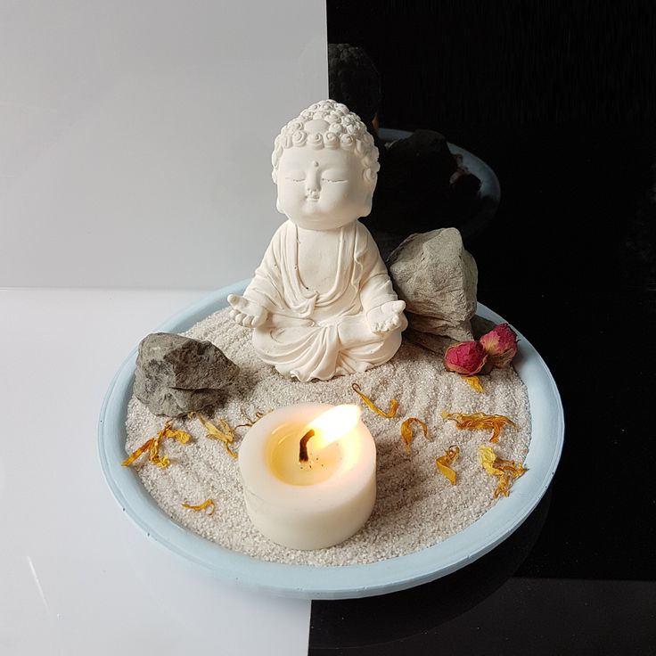 a small buddha statue sitting in the sand with a lit candle next to it on a plate