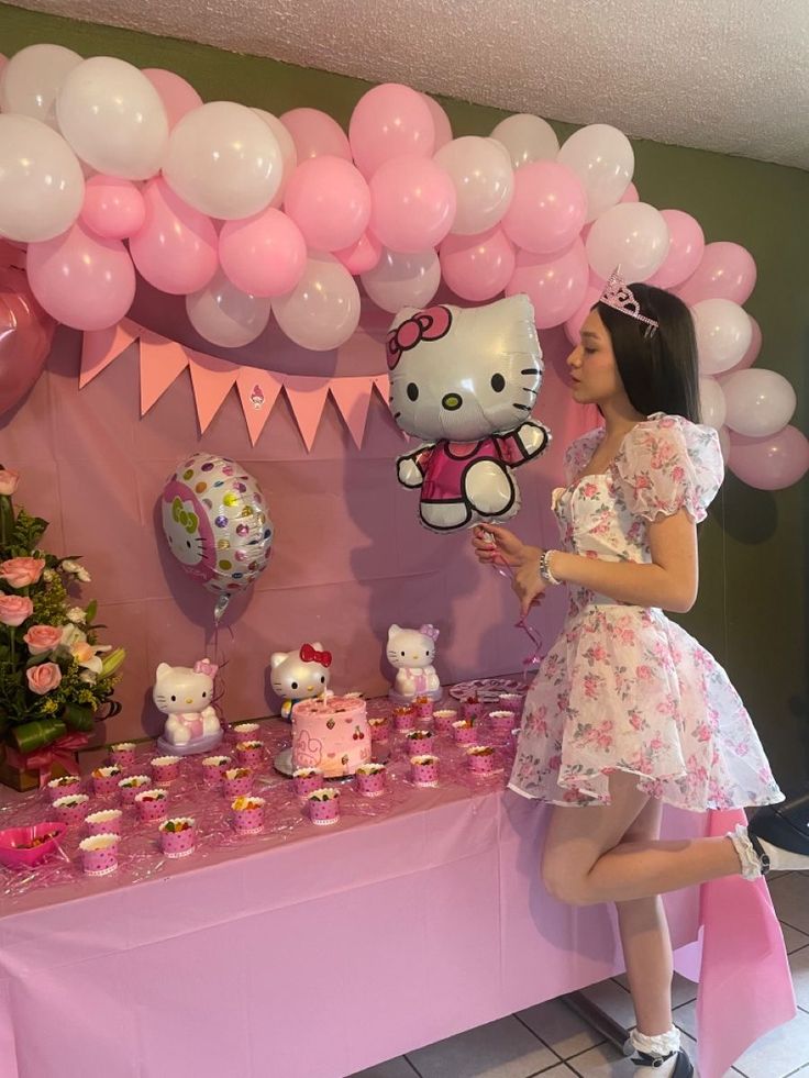 a woman standing in front of a table filled with pink and white balloons, cupcakes and hello kitty decorations