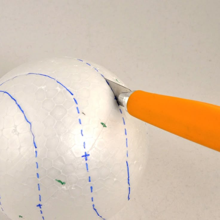 an orange handled pair of scissors cutting through a white ball with blue lines on it