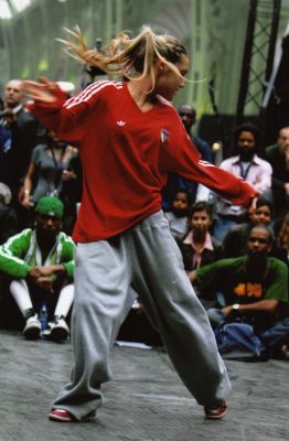 a woman in red shirt and grey pants doing a dance move on stage with people watching