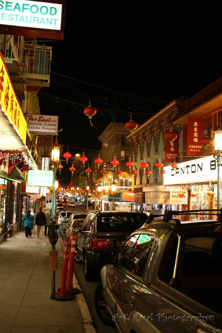 cars are parked on the street in front of restaurants and shops at night with people walking down the sidewalk