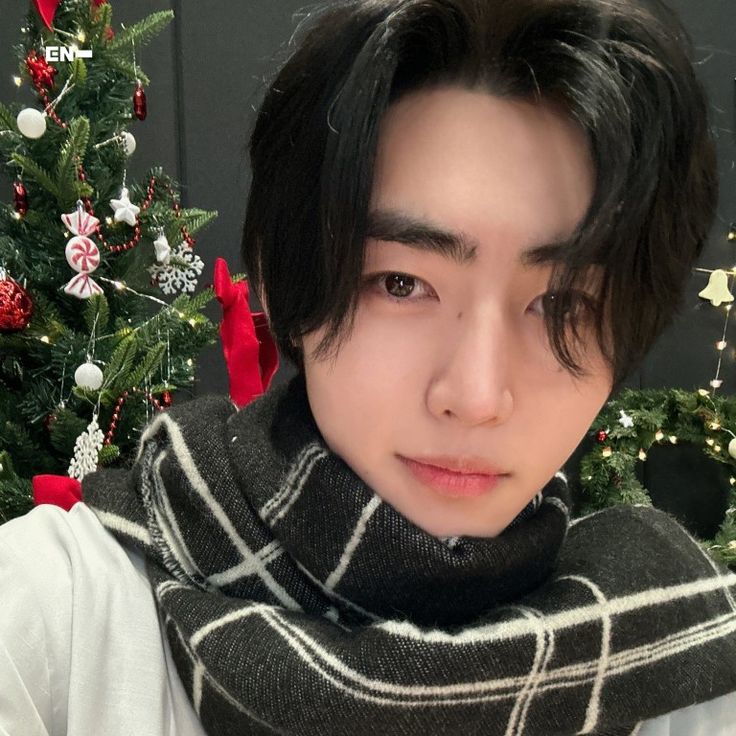 a young man with black hair wearing a scarf around his neck in front of a christmas tree