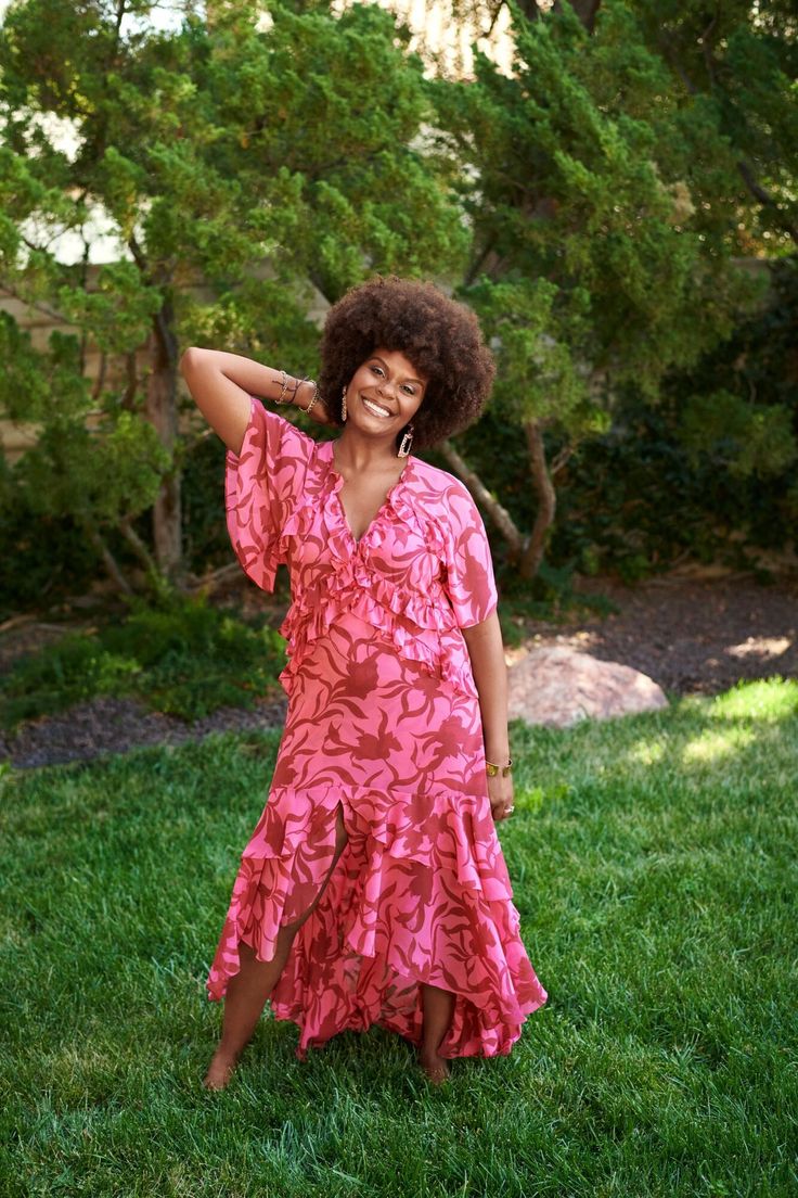 a woman standing in the grass with her arms behind her head, wearing a pink dress