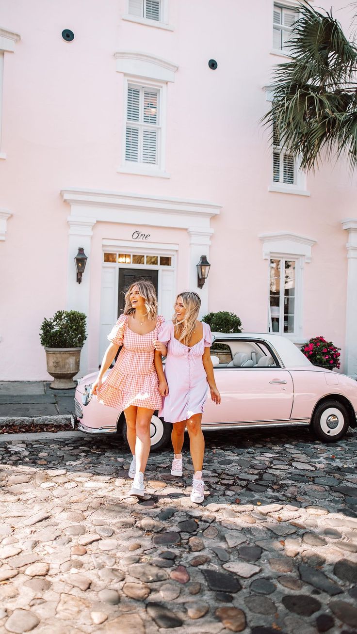 two women walking in front of a pink car on cobblestone road with palm trees