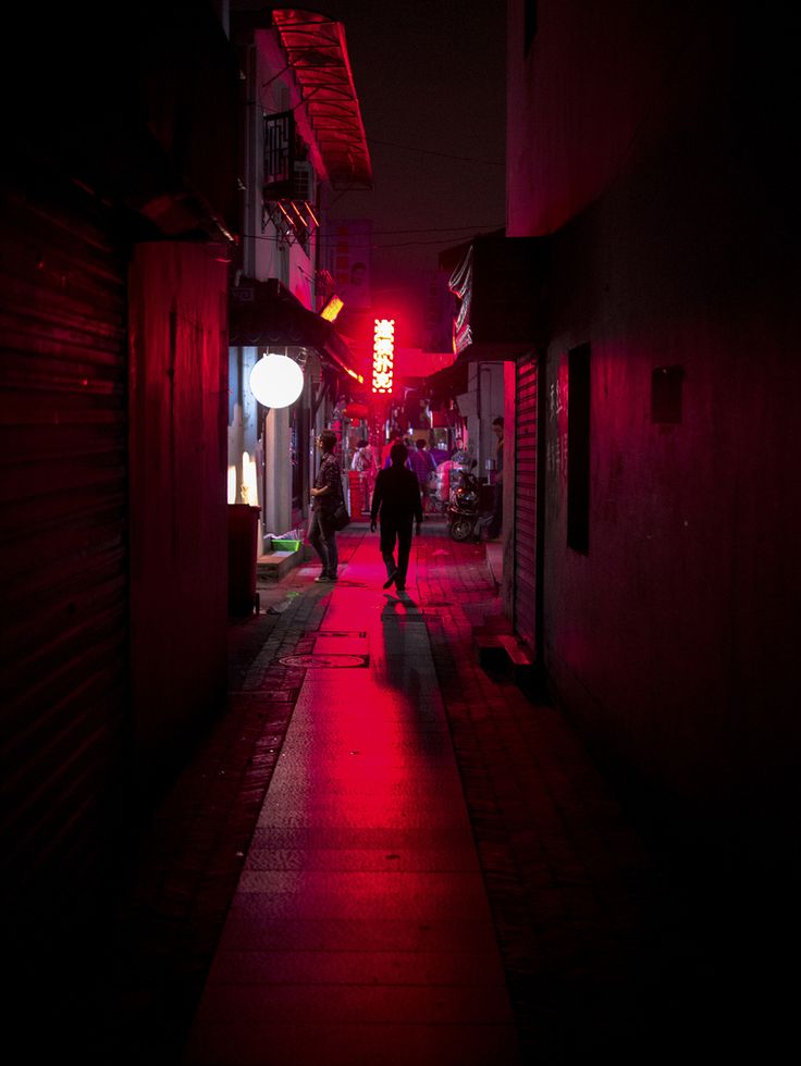 people walking down an alley way at night with red light coming from the building's windows