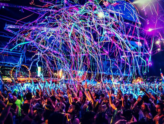 a large group of people at a concert with their hands in the air and lights on