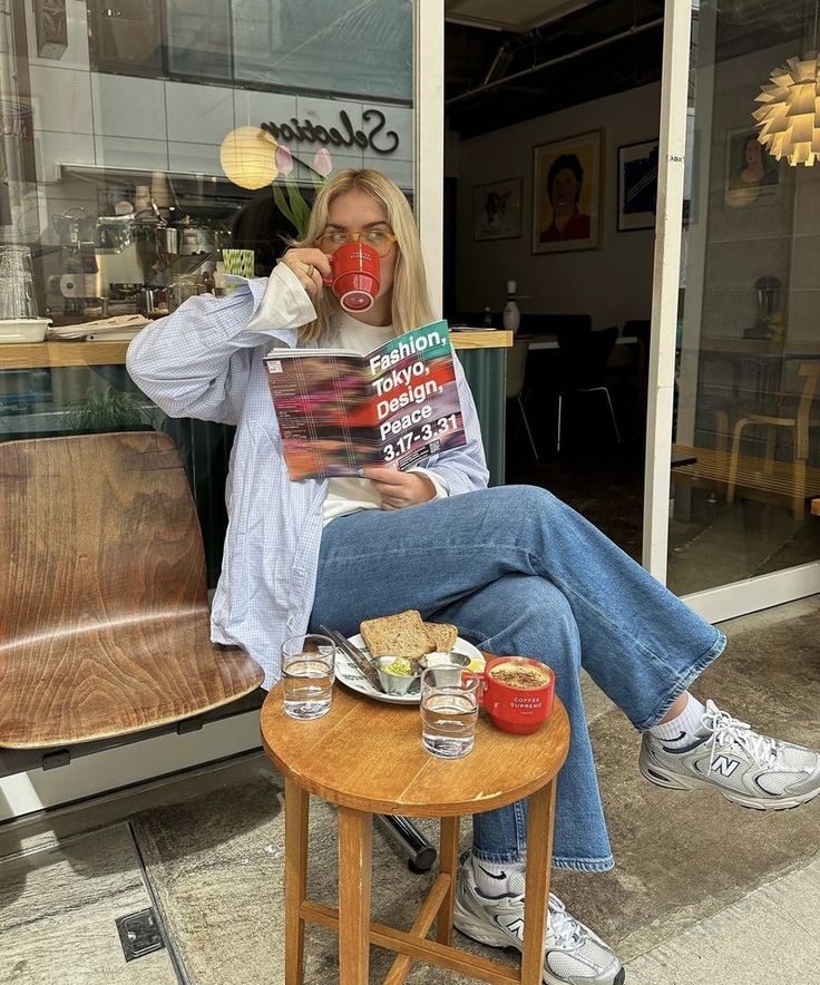 a woman sitting in a chair drinking coffee and reading a book while holding a red cup