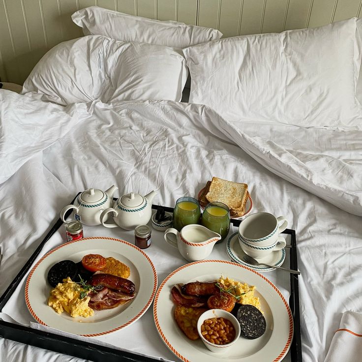 two plates with breakfast foods on them sit on a tray in front of an unmade bed