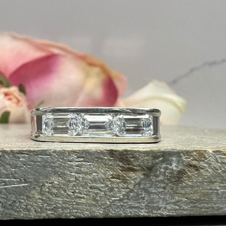 a three stone diamond ring sitting on top of a piece of wood next to a flower