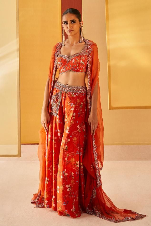 Orangish red silk organza cape with floral print and zardosi and pitta embroidery. Comes with sharara and a blouse.
Components: 3
Pattern: Printed, Embroidered
Type Of Work: Floral, Zardosi, Pitta
Neckline: Sweetheart
Sleeve Type: Cape : Flared Sleeves, Blouse : Sleeveless
Fabric: Silk organza
Color: Orange,Red
Other Details: 
Pocket detailing on sharara
Note : Outfit worn by the model on the left is not for sale.
Occasion: Mehendi and Haldi - Aza Fashions Semi-stitched Orange Sets With Motifs, Traditional Orange Embroidered Choli, Traditional Embroidered Orange Choli, Bollywood Sets With Floral Embroidery For Festive Occasions, Bollywood Festive Sets With Floral Embroidery, Bollywood Festive Set With Floral Embroidery, Orange Anarkali Sets With Intricate Embroidery, Bollywood Style Embroidered Sets With Traditional Drape, Orange Embroidered Sharara For Navratri