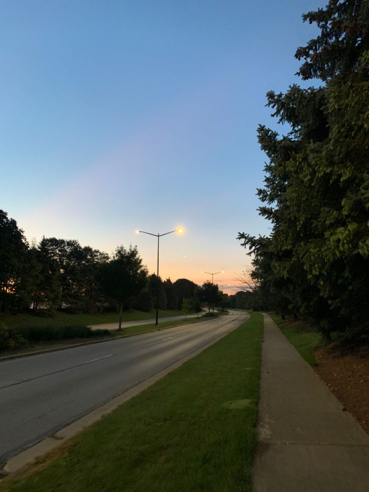 an empty street at dusk with the sun going down in the distance and trees lining the sidewalk