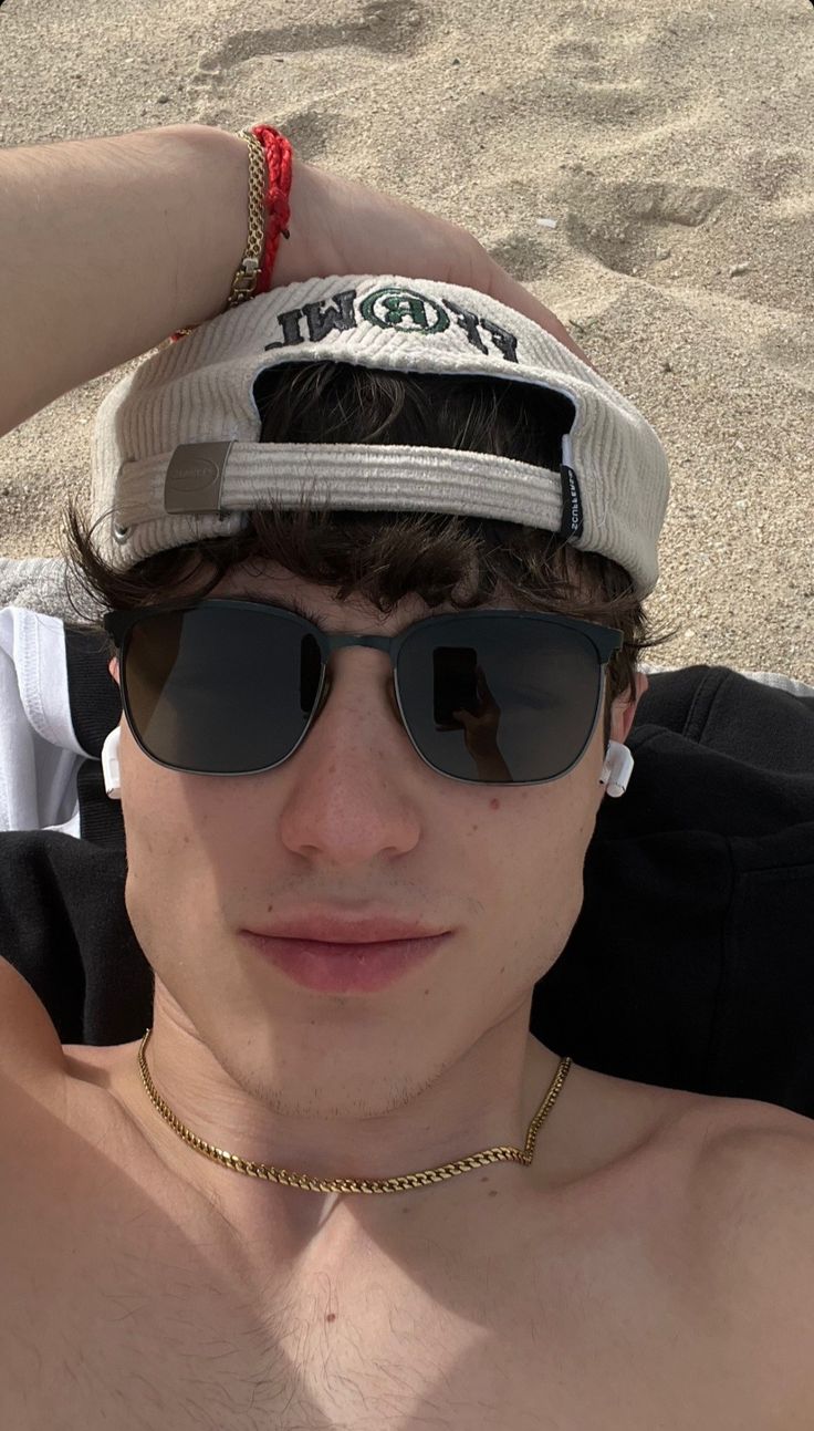 a shirtless young man wearing sunglasses and a hat on the beach with his hands behind his head