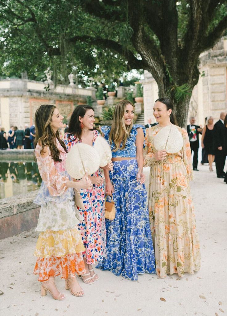 three women in dresses standing next to each other near a tree and water fountain at an outdoor event