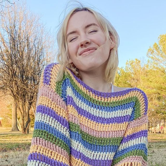 a woman wearing a multicolored crocheted sweater sitting in the grass with her eyes closed