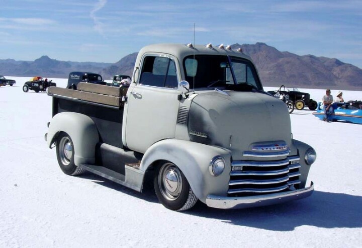 an old truck is parked in the snow with other cars behind it and people walking around