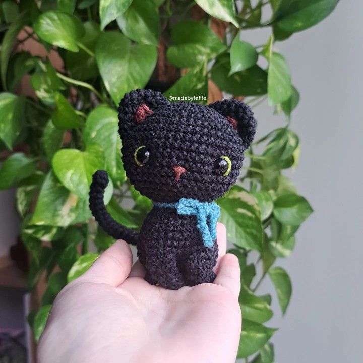 a crocheted black cat sitting on top of a hand next to a potted plant