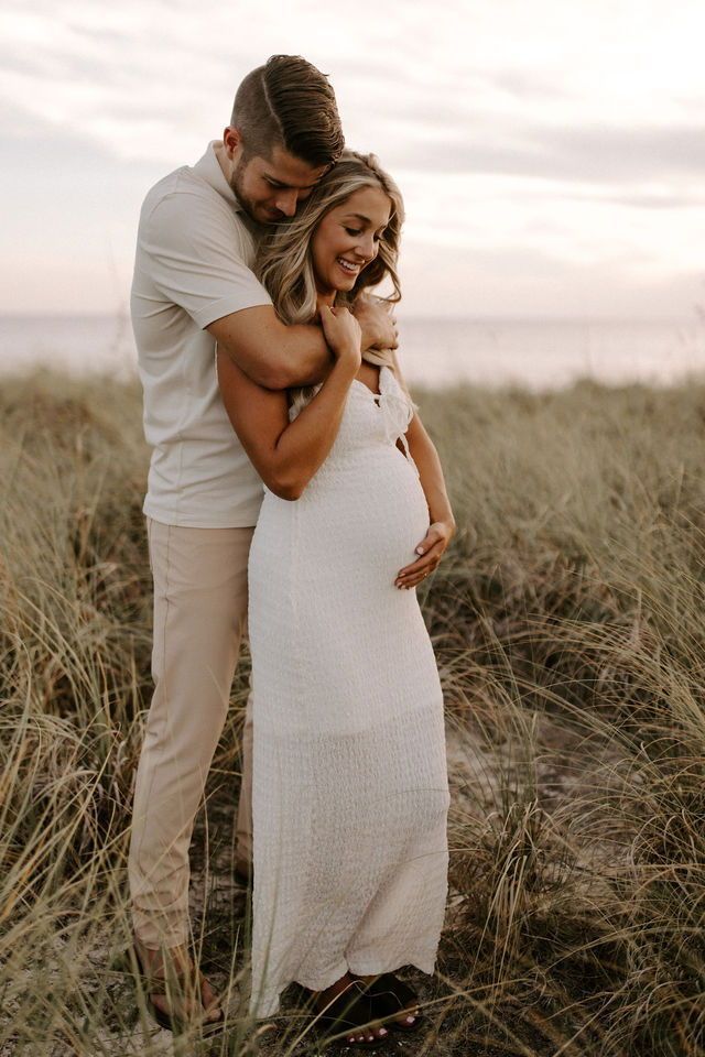 a pregnant woman standing next to a man on top of a grass covered field in front of the ocean