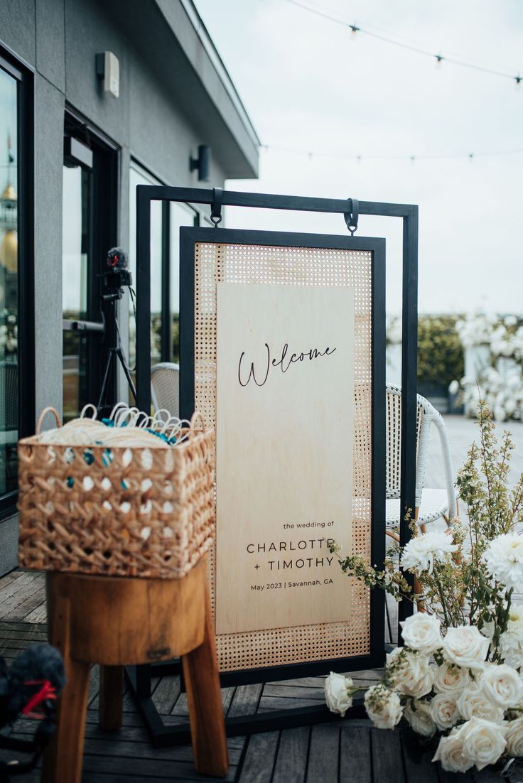 a welcome sign sitting on top of a wooden table next to flowers and baskets in front of a building