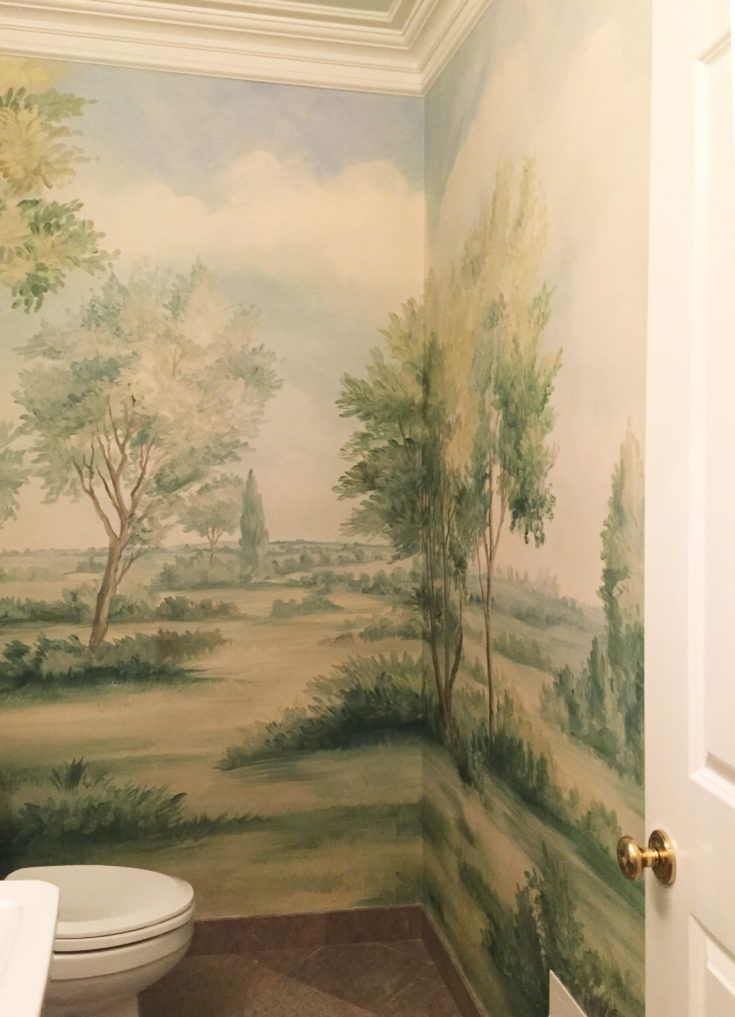 a toilet in a bathroom next to a wall with trees painted on it and a sky background