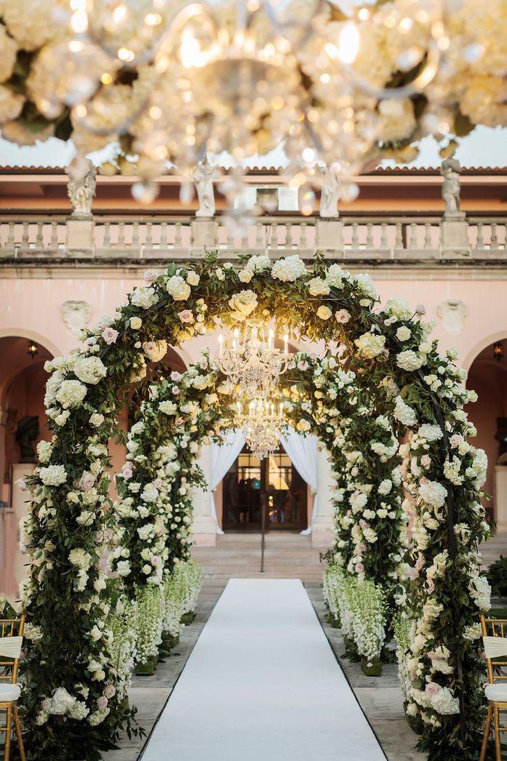 an archway decorated with white flowers and greenery for a wedding ceremony in front of a chandelier