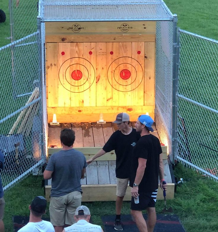 three men standing in front of a wooden box with darts on it and people watching