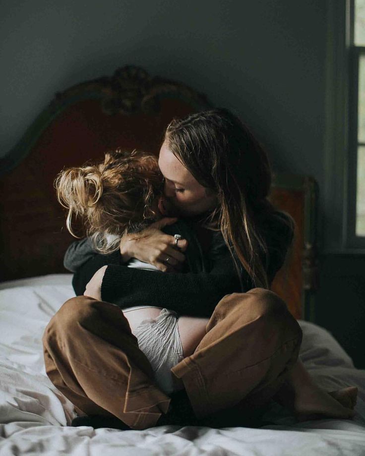 a woman sitting on top of a bed hugging another woman's head in her arms