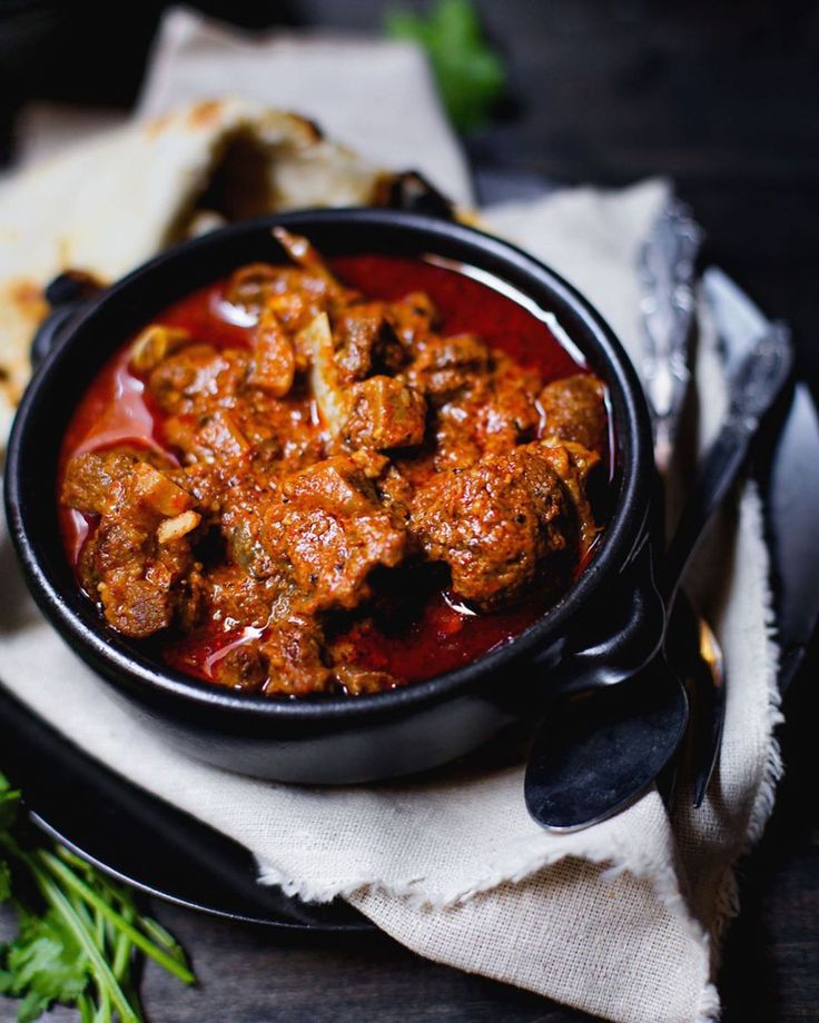 a black bowl filled with meat and sauce on top of a white napkin next to bread