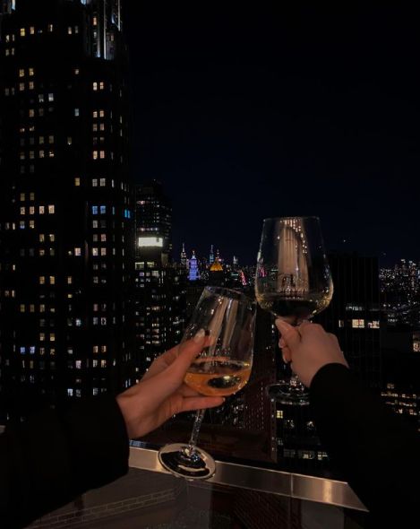 two people toasting wine glasses on top of a building at night with city lights in the background