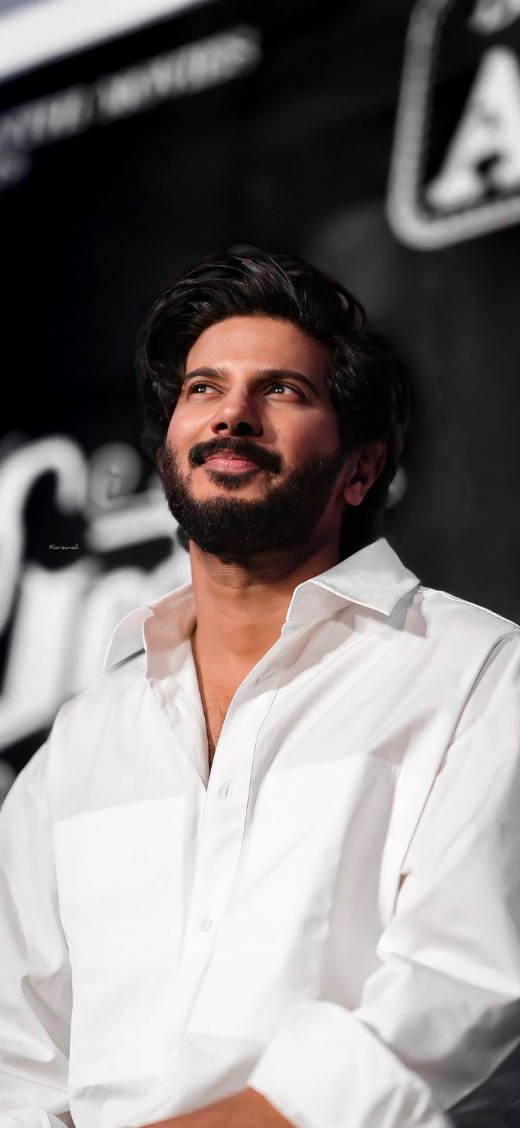 a man with a beard sitting in front of a black and white background wearing a white shirt