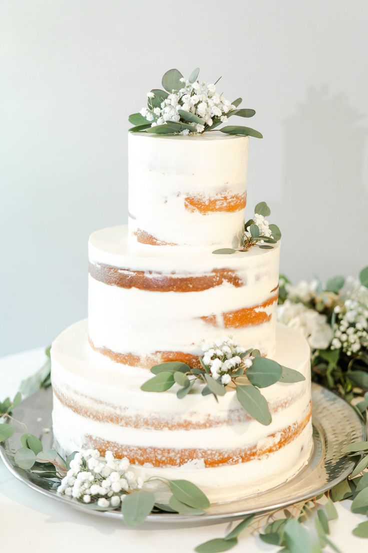 a three tiered white cake with greenery on the top and bottom, sitting on a silver platter