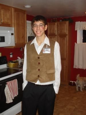 a young man standing in a kitchen wearing a brown vest and white shirt with his hands in his pockets