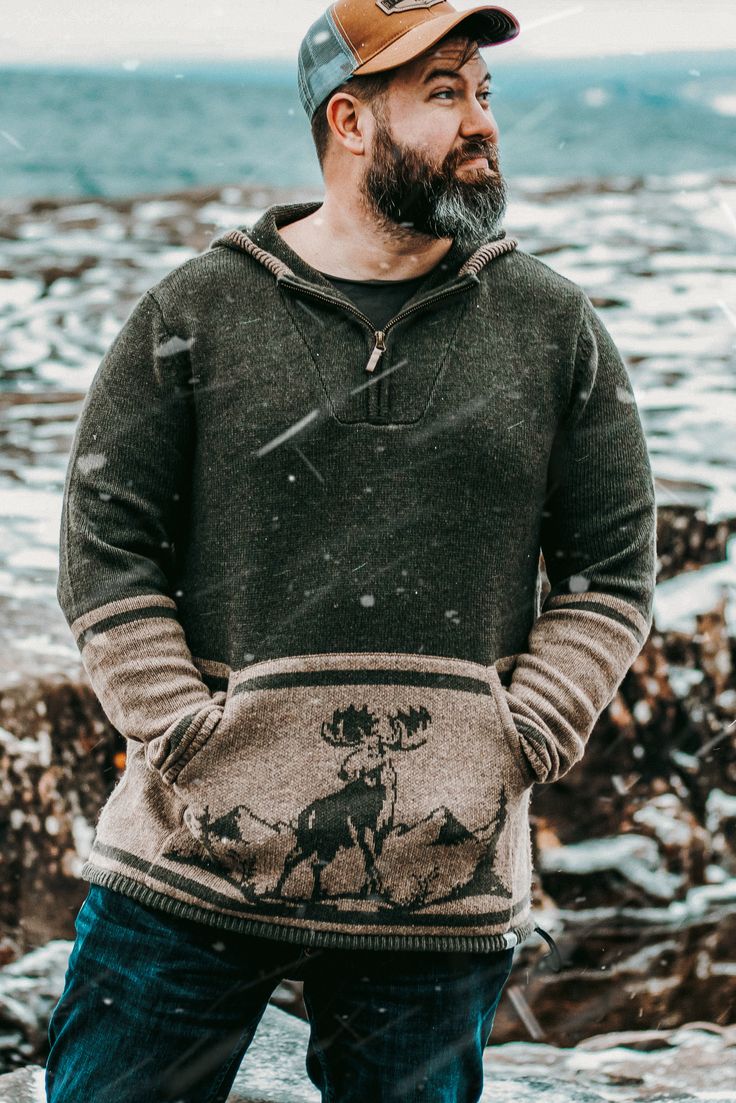 a man standing on the beach wearing a sweater with an elk print and a hat