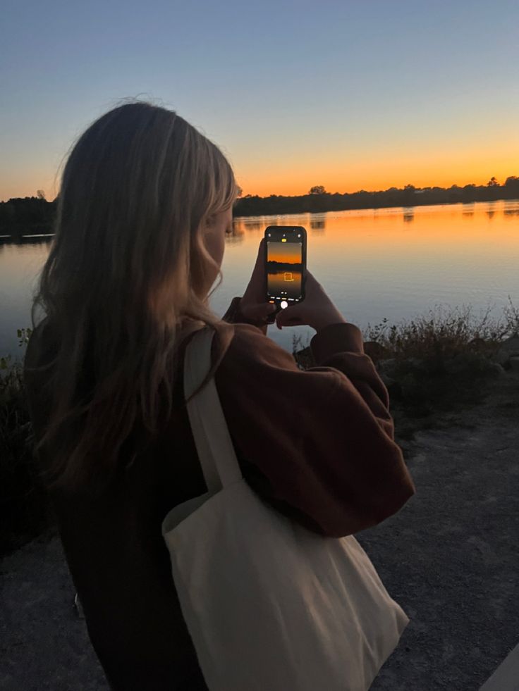 a woman is taking a photo with her cell phone at sunset by the water's edge