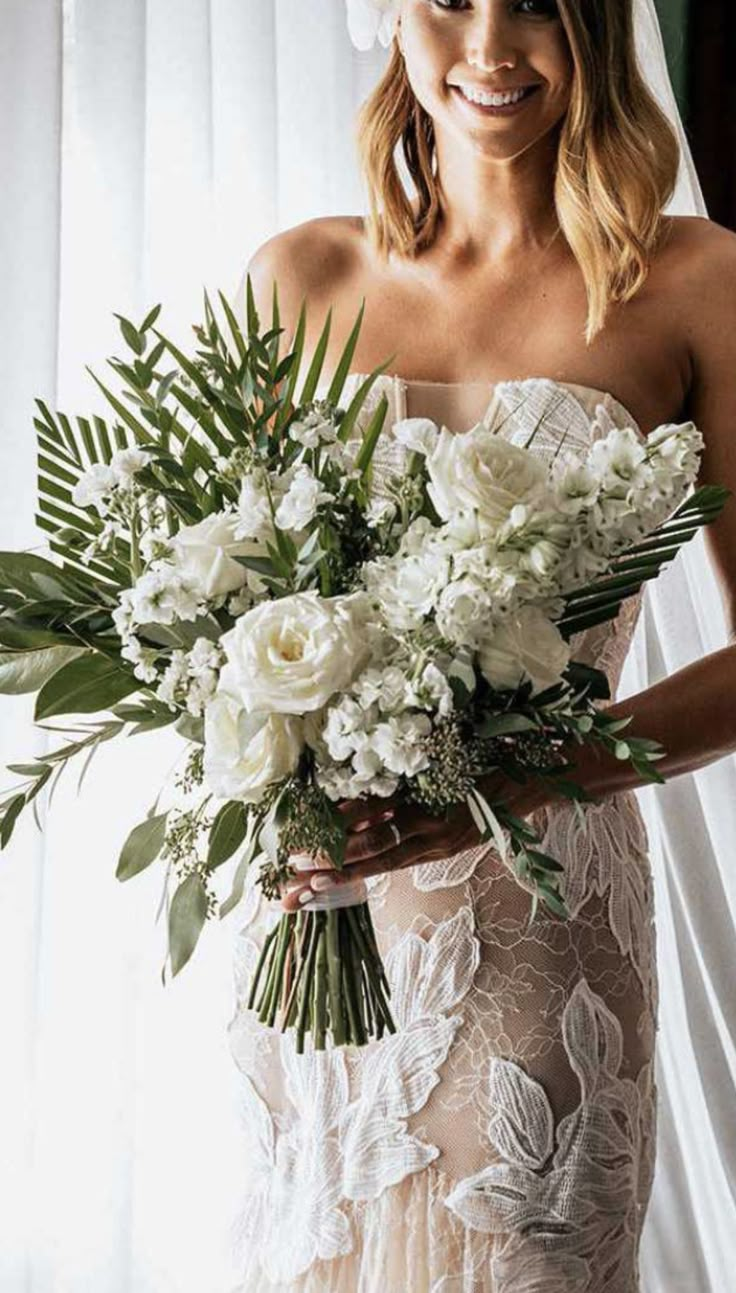 a woman in a wedding dress holding a bouquet of white flowers and greenery, smiling at the camera