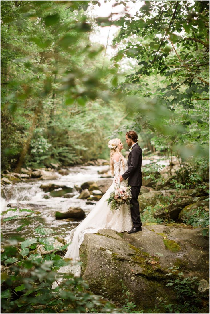 a bride and groom standing on top of a rock in the woods next to a river