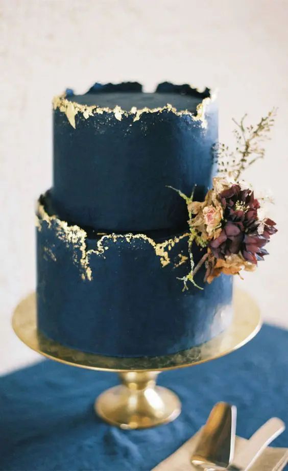 a three tiered blue cake with gold sprinkles and flowers on top