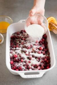 a person pouring sugar into a dish full of cranberries and orange juices