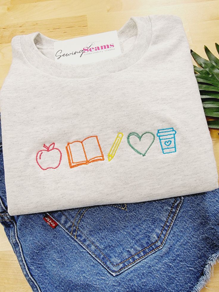 Brighten a teacher’s day with our Teacher Elements Embroidered T-Shirt! This delightful t-shirt features an array of teacher icons embroidered in the vibrant colors of the rainbow, creating a simple yet effective design that celebrates the passion and dedication of educators. Crafted from high-quality, soft, and breathable fabric, this t-shirt offers all-day comfort and durability. It’s perfect for wearing to school, casual outings, or lounging at home, making it a versatile addition to any teac Cute Teacher Sweatshirts, Teacher Shirt Designs Ideas, Teacher Hand Embroidery, Embroidery For Teachers Gift Ideas, Embroidered Teacher Gifts, Simple Tshirt Design Ideas, Machine Embroidery Gift Ideas, Class Shirt Ideas, Teacher T Shirts Ideas Design