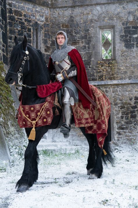 a man dressed as a knight riding on the back of a black horse in front of a stone building