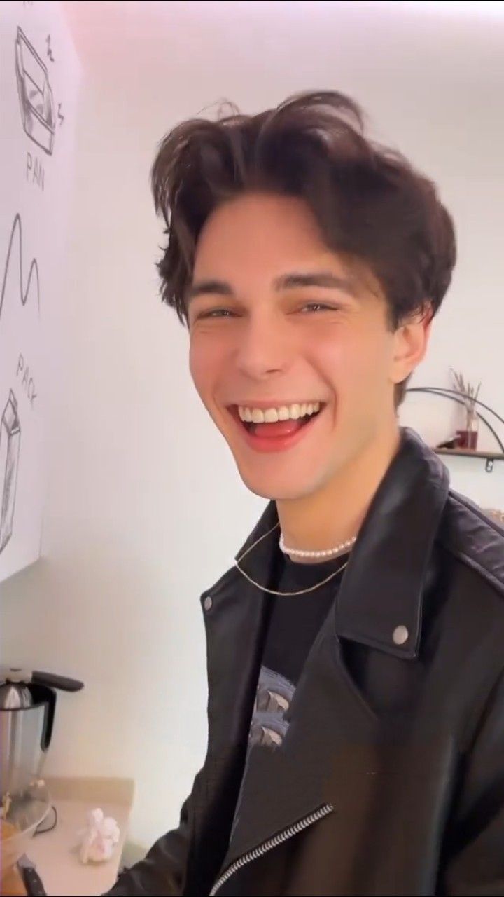 a young man wearing a black jacket and smiling at the camera while sitting in front of a whiteboard