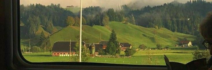 a woman sitting on a train looking out the window at green hills and houses in the distance