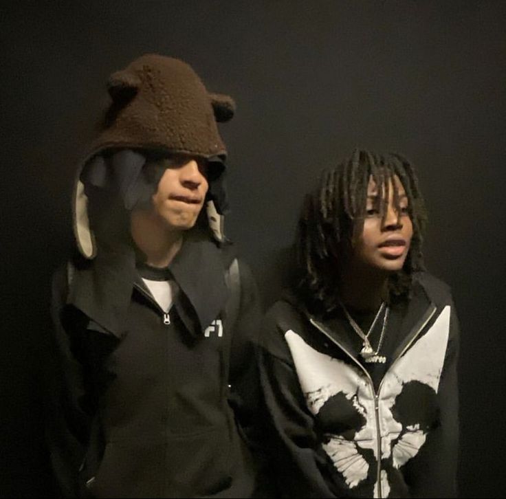 two young men standing next to each other in front of a black wall wearing hoodies