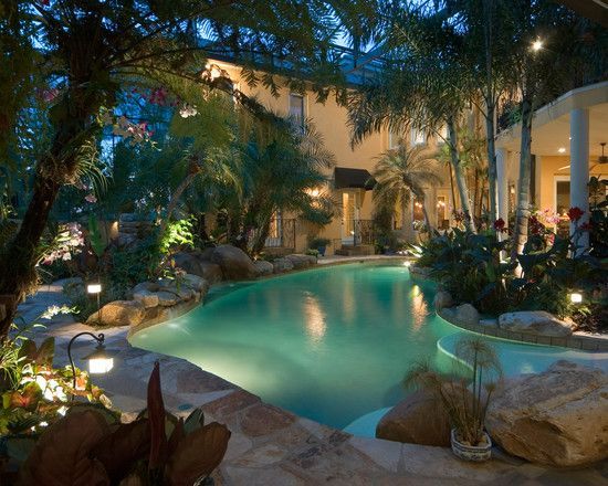 an outdoor swimming pool surrounded by trees and rocks at night time with lights on the water